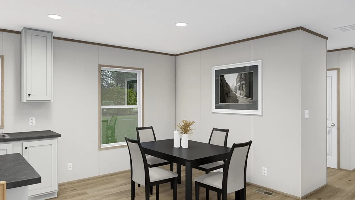 A small dining area with a black table, four chairs, a vase with dried flowers, a wall-mounted photograph, and two windows in a room with light-colored walls and wood flooring.