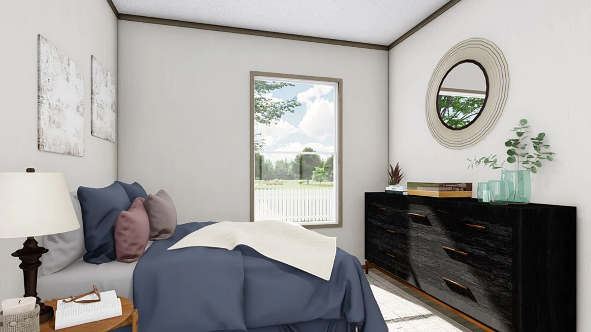 A bedroom with a bed covered in blue bedding, a black dresser with decor items, a round mirror on the wall, and a window showing an outdoor view.