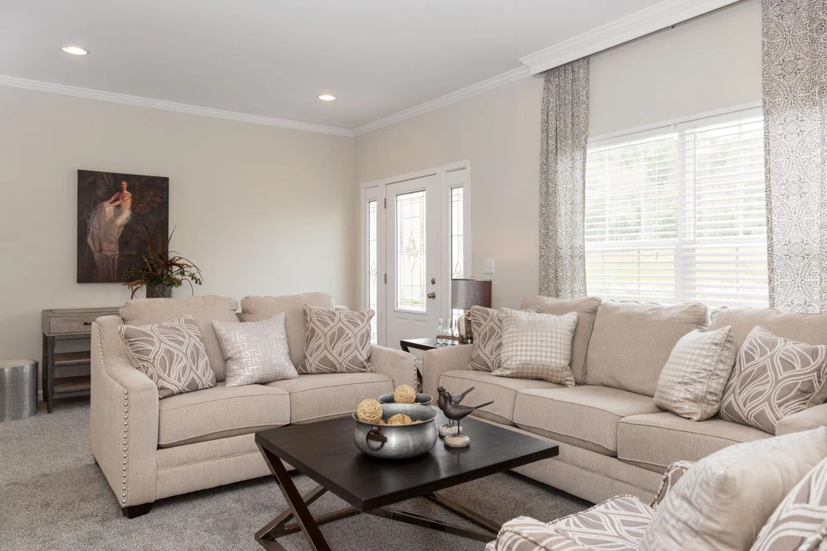 A living room with beige sofas, patterned pillows, a dark coffee table with decorative items, light walls, large windows with curtains, and a painting on the wall.