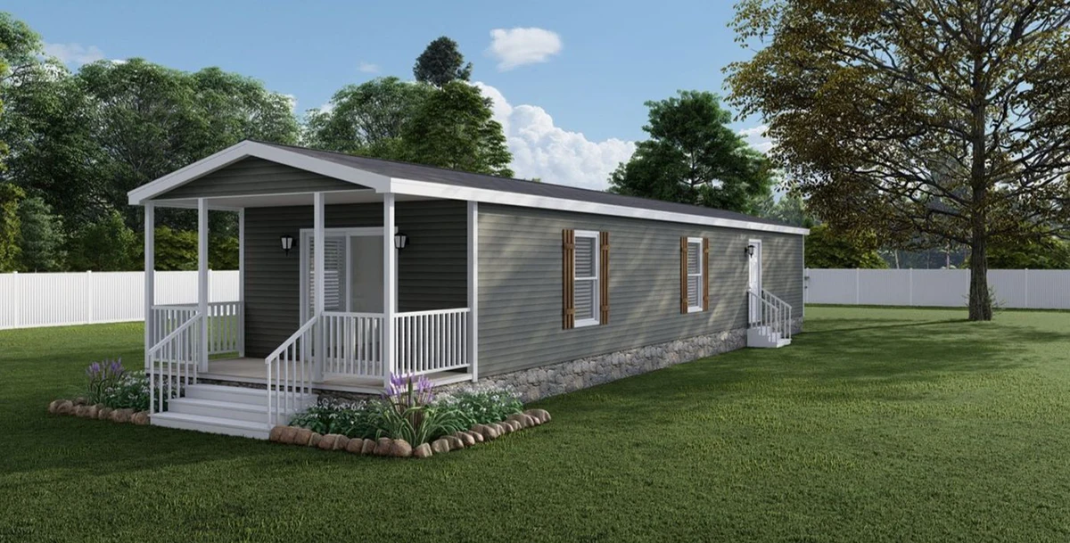 A single-story gray manufactured home with a small covered porch, white trim, and landscaped garden beds, set in a grassy yard with trees and a white fence.
