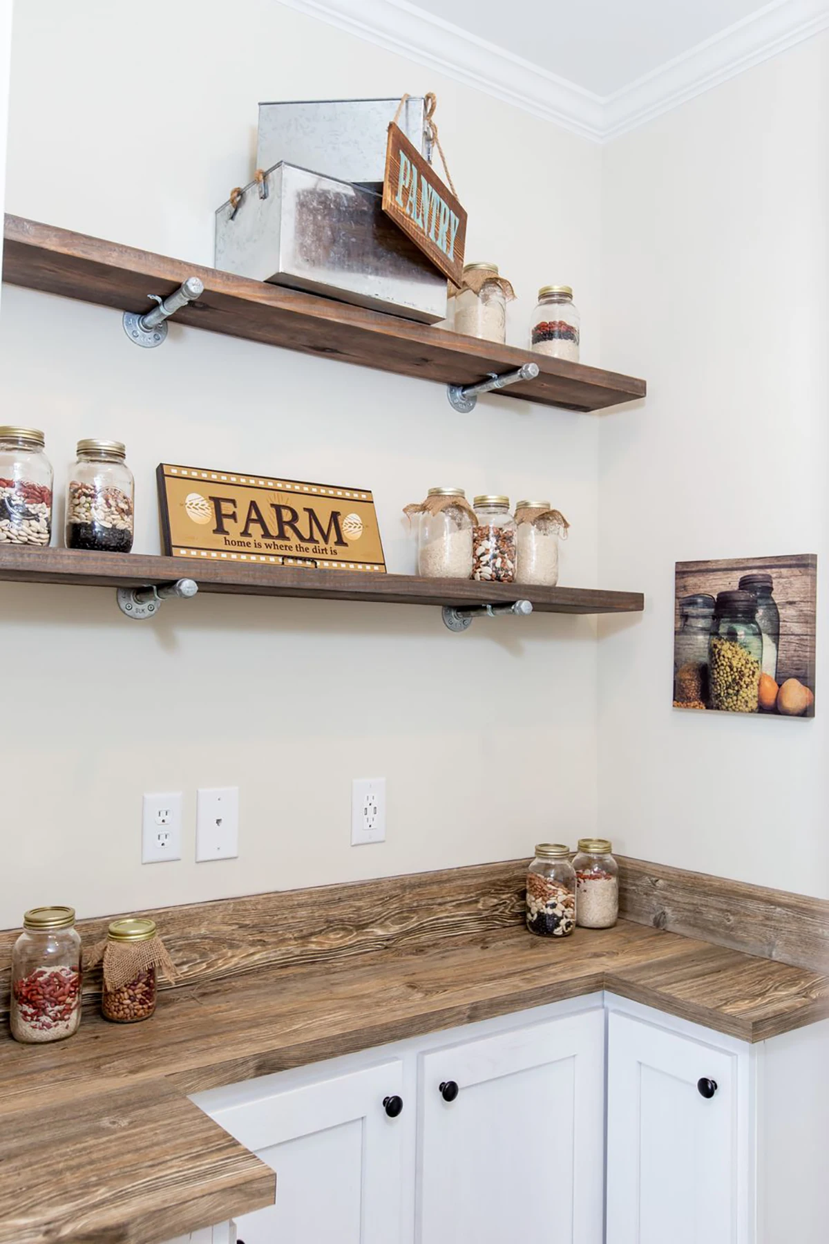 A small pantry corner with wooden shelves holding glass jars of dry goods and decorative farm-themed signs, with white cabinets and a rustic wood countertop below.