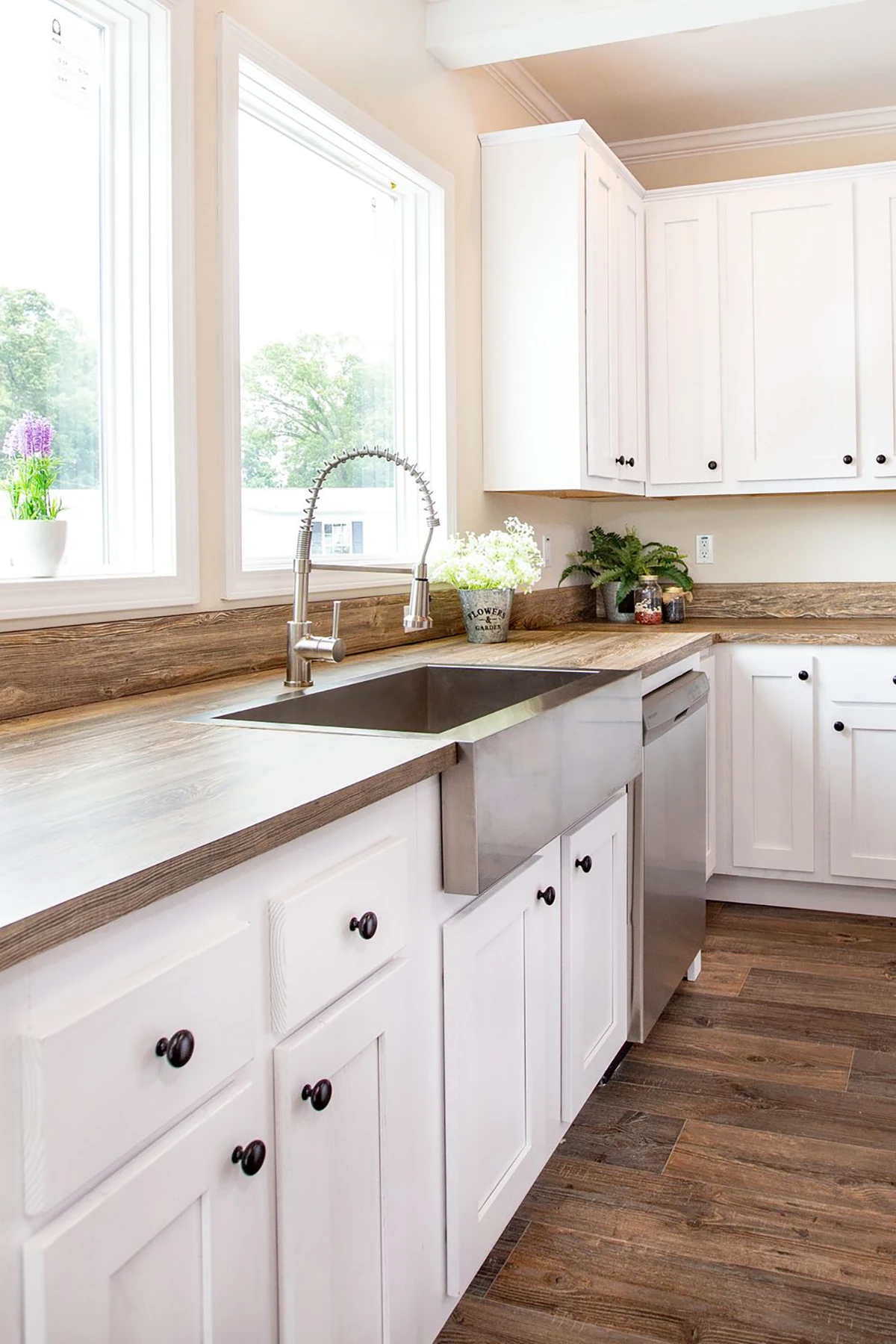 Modern kitchen with white cabinets, stainless steel farmhouse sink, dishwasher, wood countertops, and potted plants by large windows.