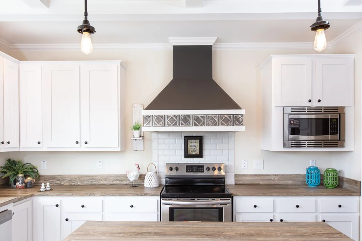 Modern kitchen with white cabinets, stainless steel oven, black range hood, microwave, pendant lights, and decorative items on the countertop.