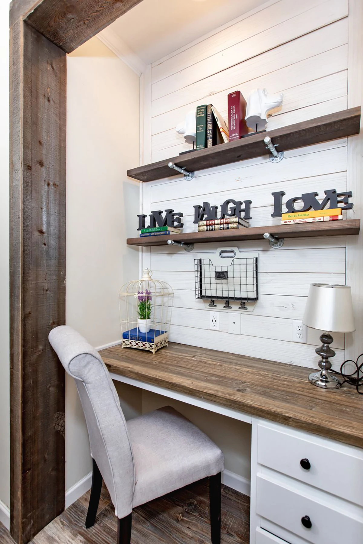 A small home office nook with a rustic wood desk, white chair, shelves holding books and decor, and a lamp on the desk. A caged plant and "Live Laugh Love" signs are displayed.