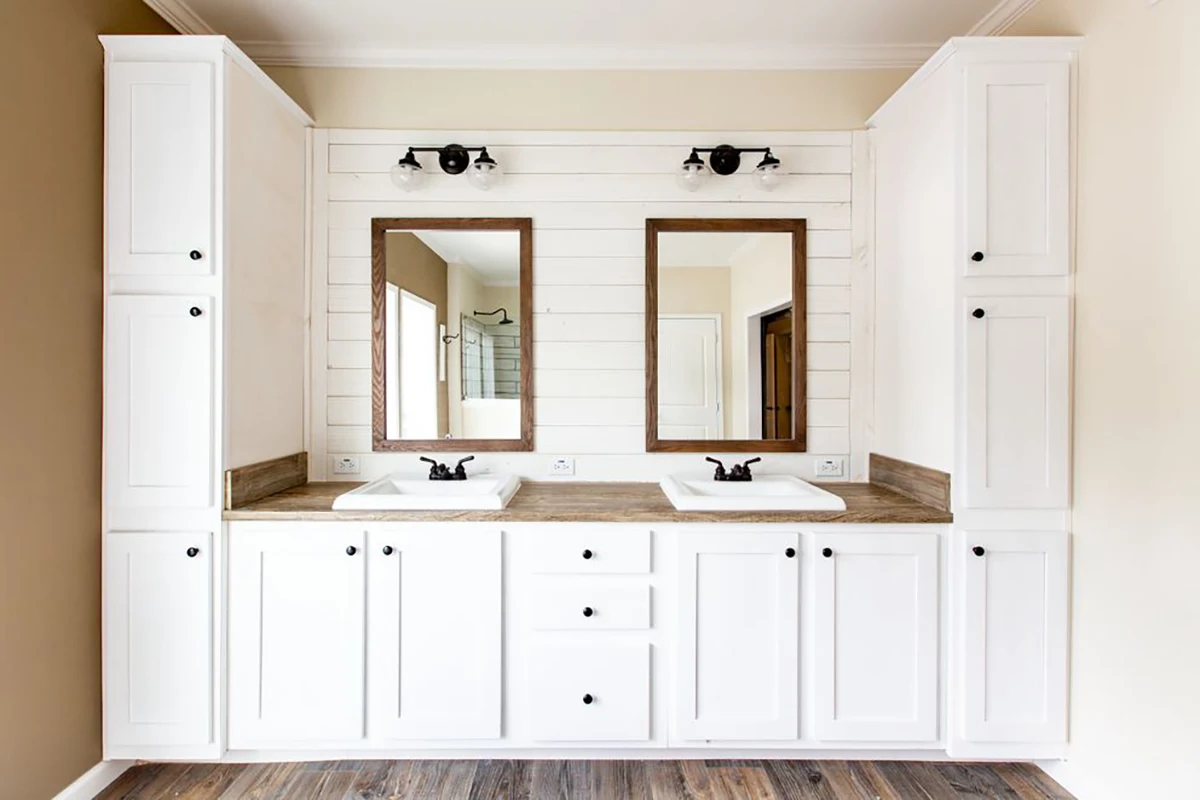 White double bathroom vanity with wooden countertop, two sinks, two framed mirrors, wall-mounted lights, and storage cabinets, set against a white shiplap wall.