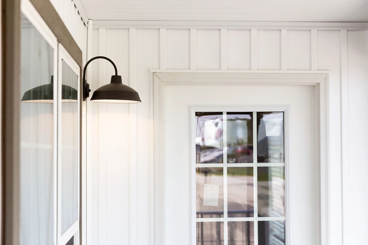 A black outdoor wall sconce light is mounted next to a white framed door with window panes on a white panel wall.