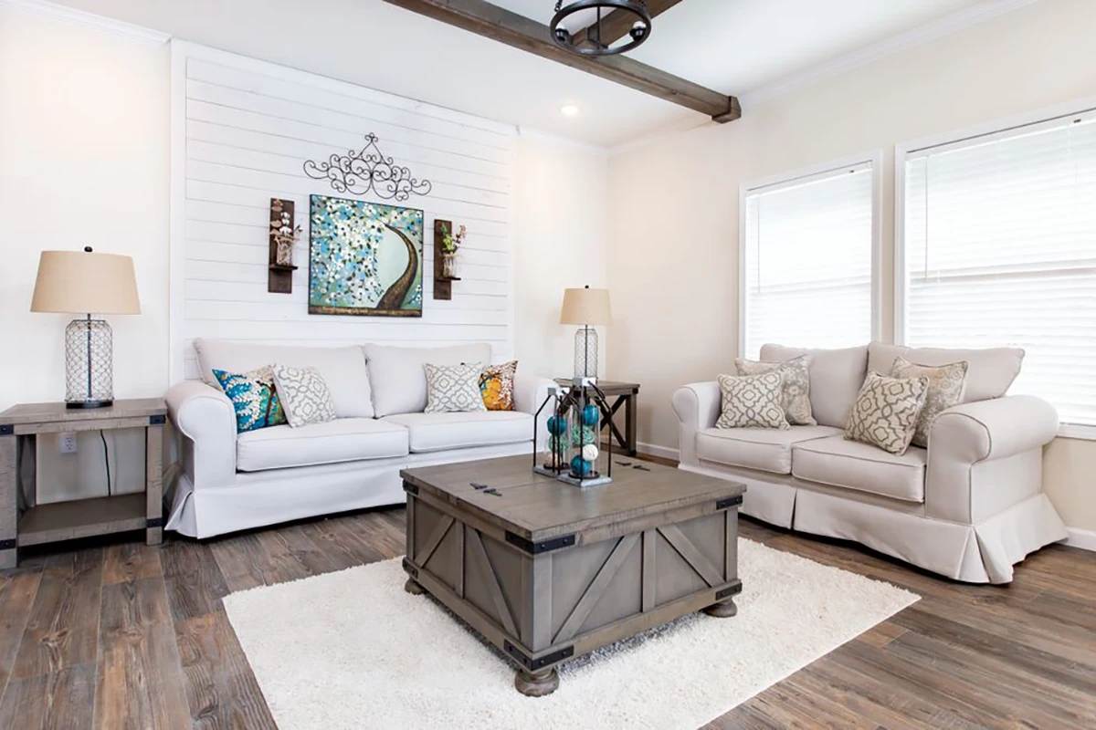 A bright living room with two beige sofas, patterned cushions, a wooden coffee table, side tables with lamps, wall art, and hardwood flooring.