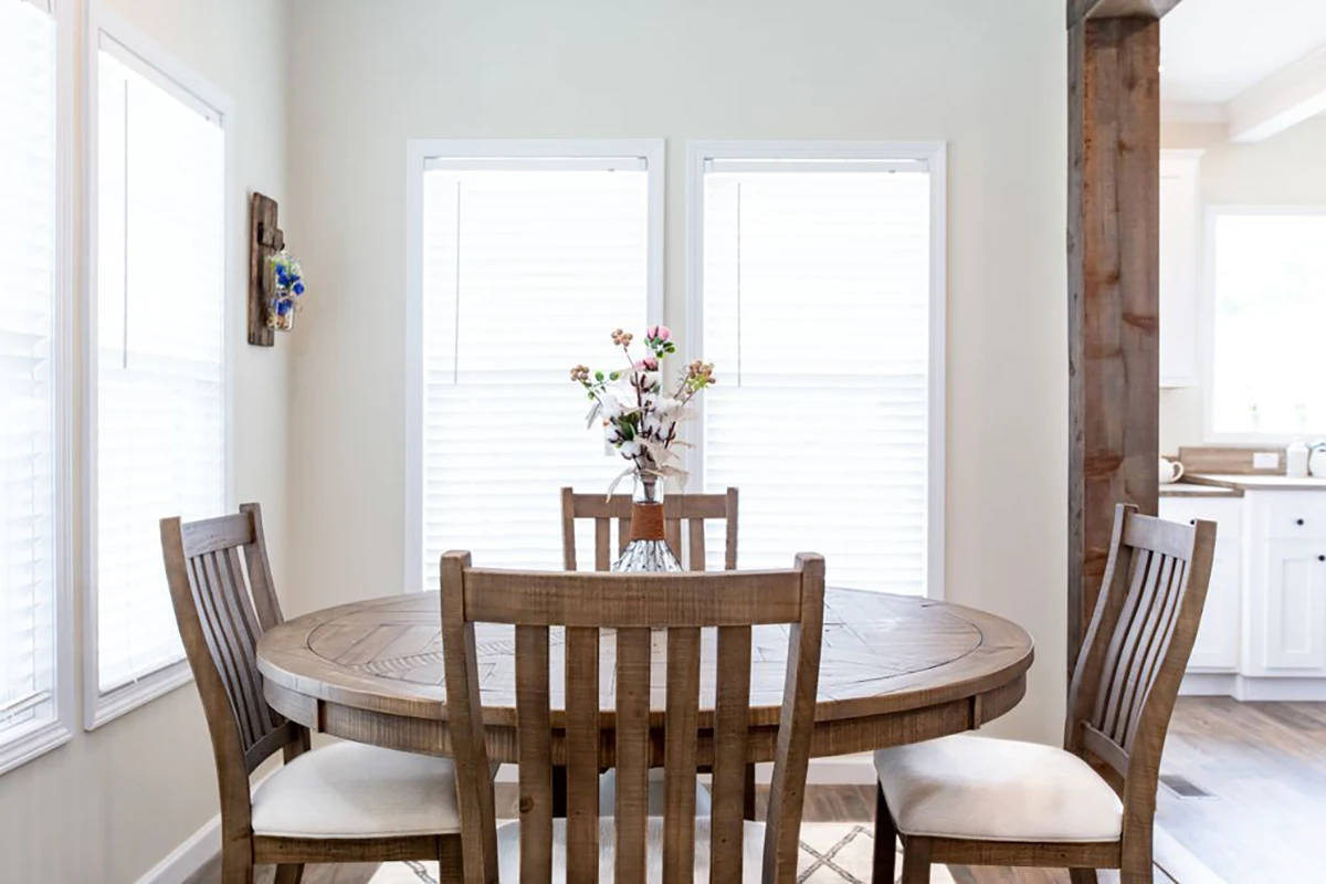 A round wooden dining table with four matching chairs and a vase of flowers, set in a bright room with two large windows and white walls.
