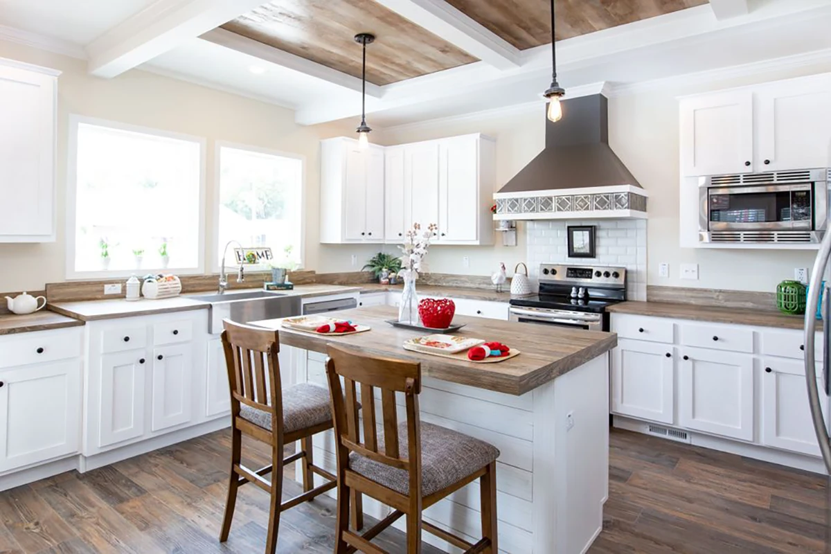 Modern kitchen with white cabinets, wood accents, an island with two chairs, stainless steel appliances, and large windows letting in natural light.