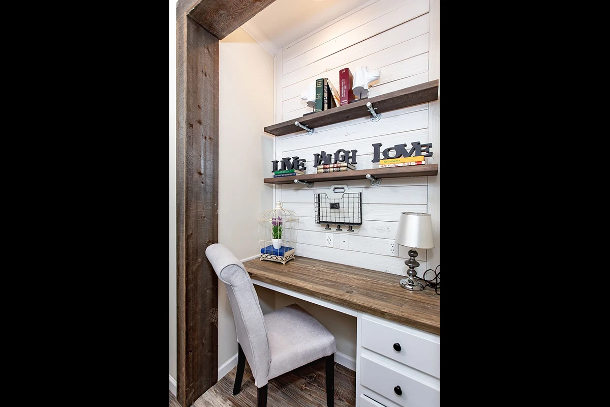 Southern Charm 13 Small home office nook with a wooden desk, upholstered chair, two shelves displaying books and decor, lamp, potted plant, and a wire organizer mounted on a white shiplap wall.