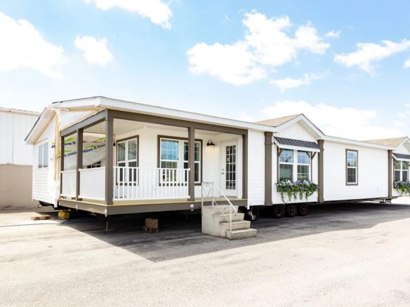 A large white manufactured home with a covered porch and steps, set on a paved lot under a partly cloudy sky.
