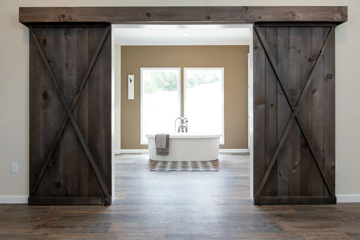 Southern Charm 6 A white freestanding bathtub is centered in front of two large windows, framed by open dark wooden barn doors on a wood floor.