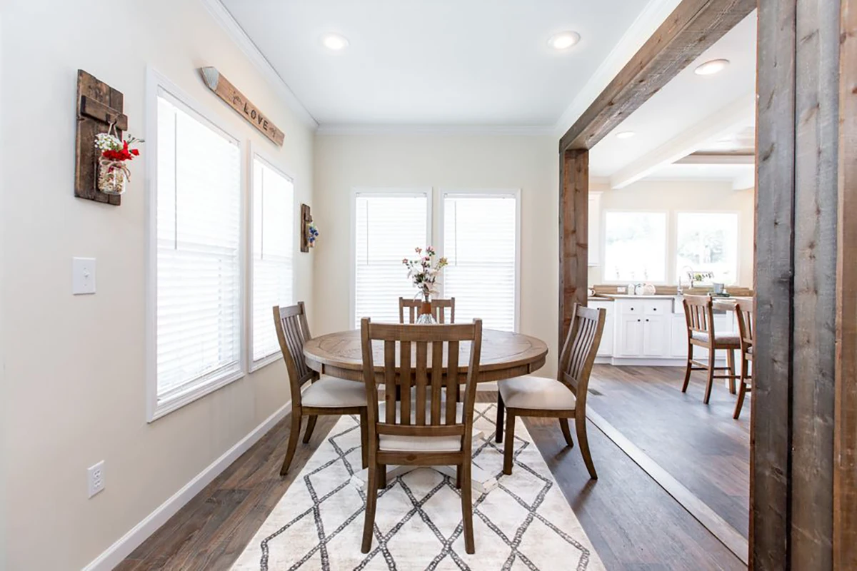 Southern Charm 9 Bright dining area with a round wooden table, four chairs, and a centerpiece. Large windows provide natural light. Kitchen with white cabinets visible in the background.