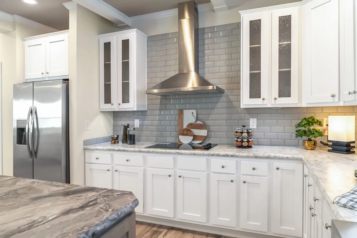 Modern kitchen with white cabinets, stainless steel appliances, gray subway tile backsplash, marble countertop, and a central range hood. Decorative items and a lamp are on the counter.