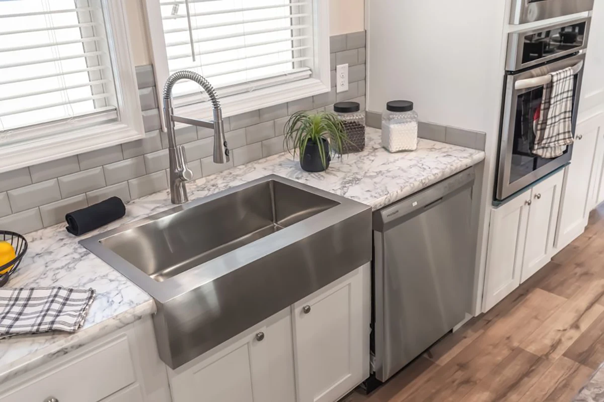 Modern kitchen with a stainless steel farmhouse sink, marble countertop, dishwasher, double ovens, and jars on the counter.