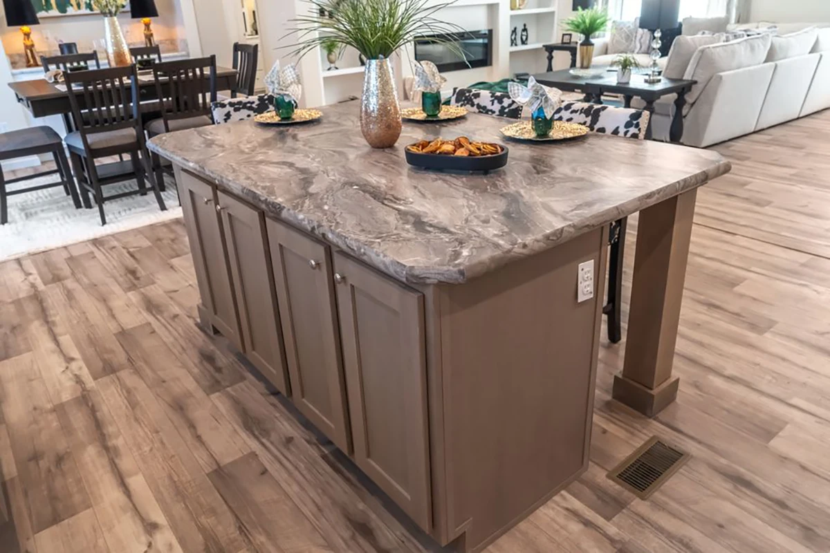 A kitchen island with a gray marble countertop, brown cabinets, and place settings for four, situated in an open-plan dining and living area with wood flooring.