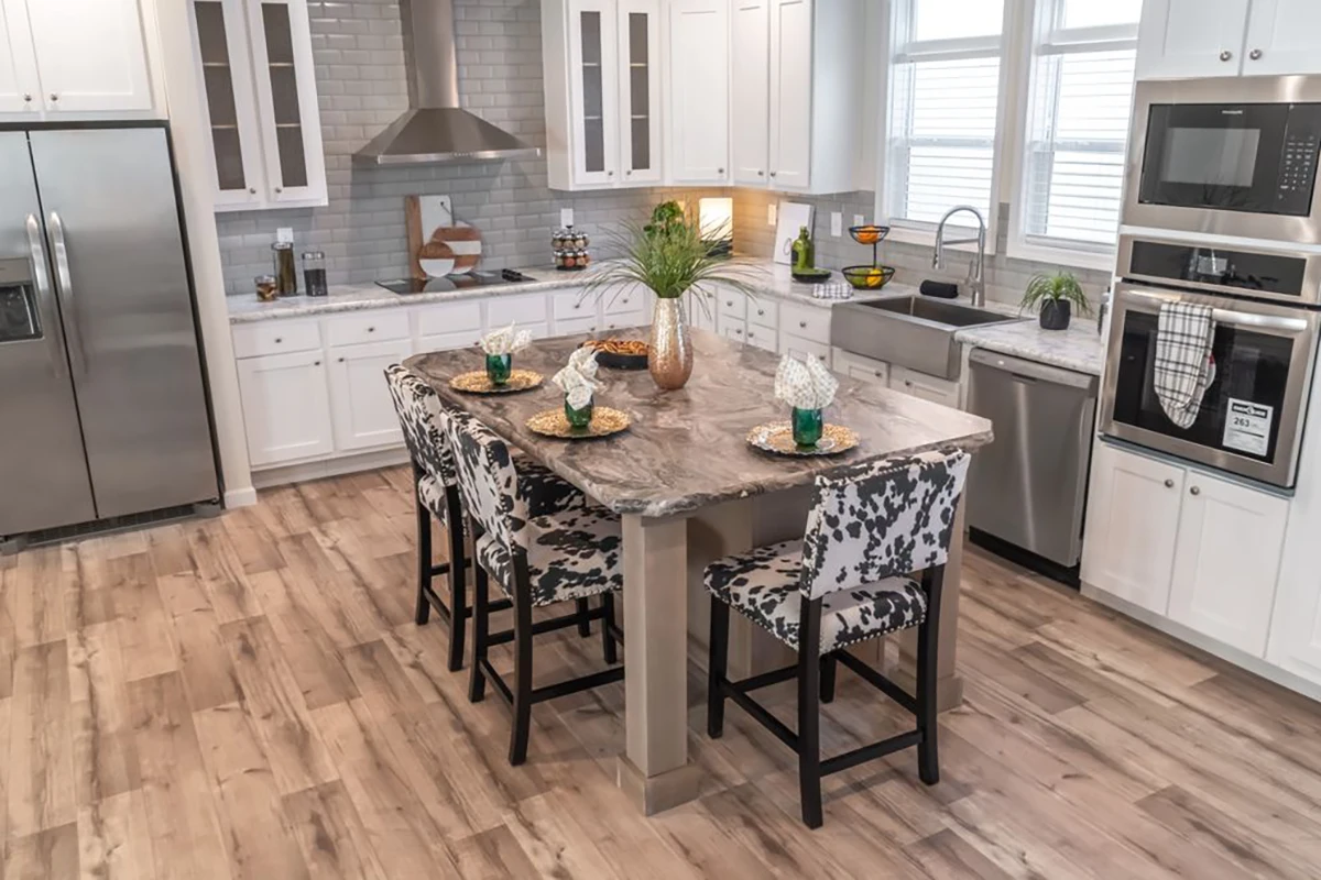 Modern kitchen with white cabinets, stainless steel appliances, a marble island, and four patterned chairs. Light wood flooring and large windows brighten the space.