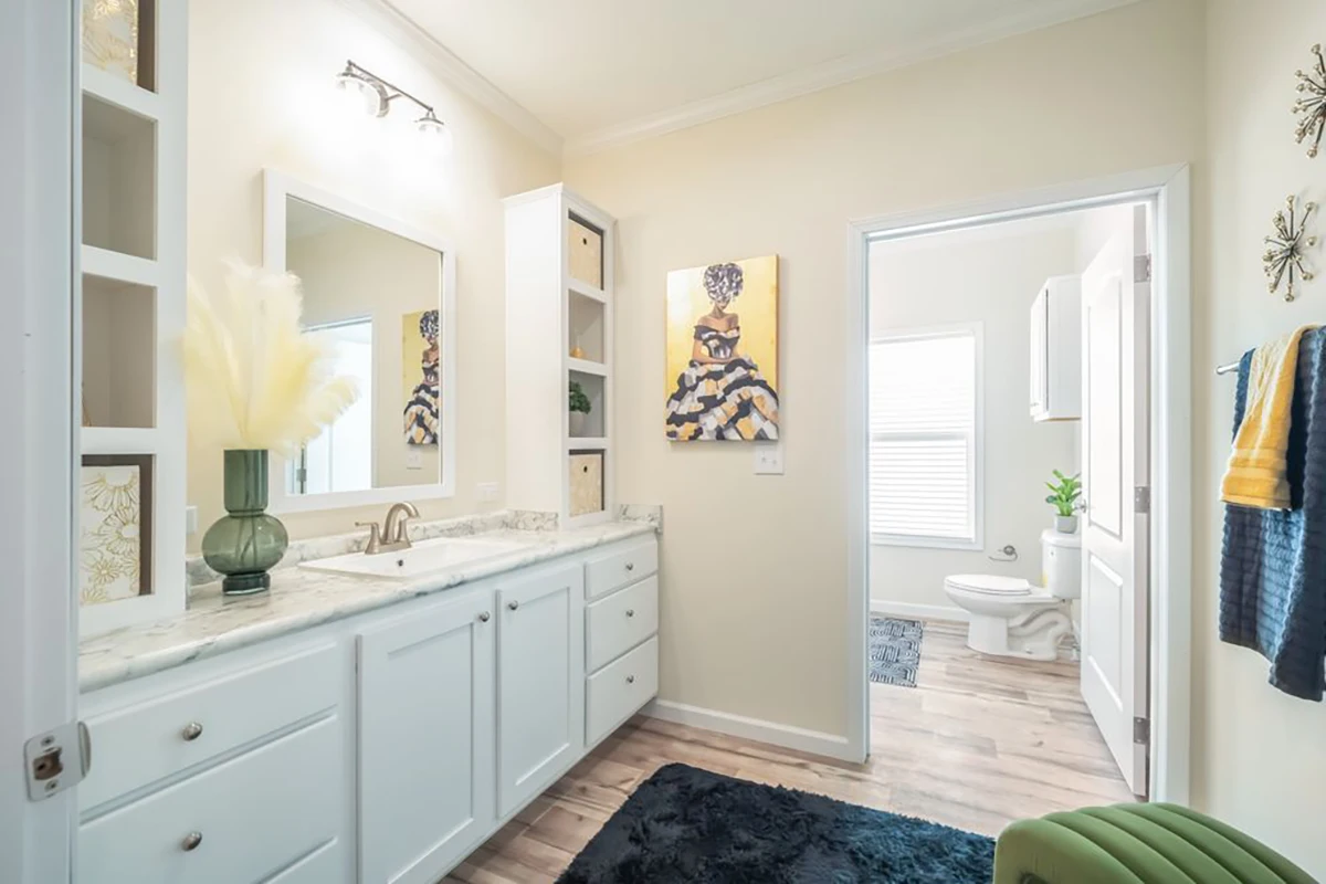 Bright bathroom with white cabinets, marble countertop, wall art above a black rug, and a view into a separate toilet area with window and greenery.