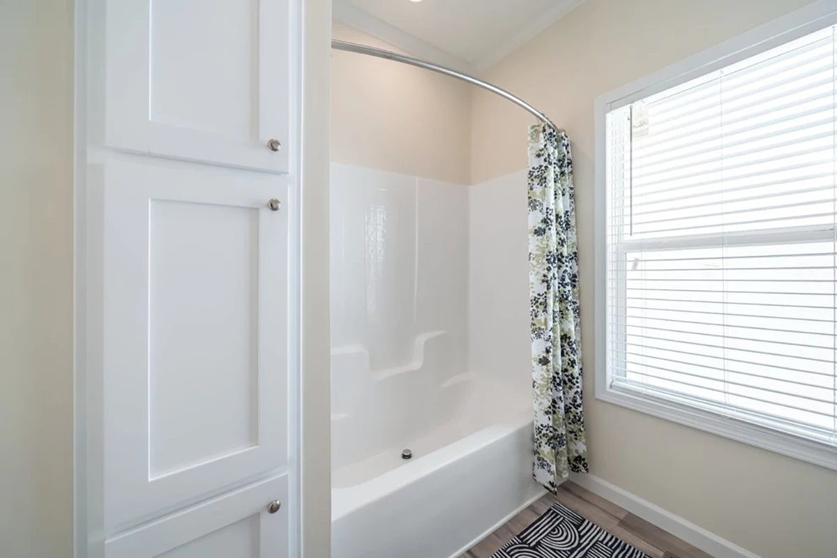 White bathroom with a built-in bathtub and shower, floral shower curtain, white storage cabinets, window with blinds, and a patterned rug on the floor.