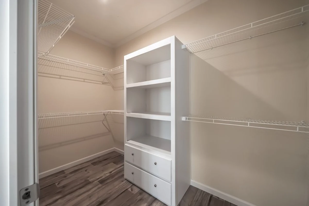 Empty walk-in closet with wire shelving on three walls and a white built-in shelving unit with drawers in the center, featuring wood-look flooring and beige walls.