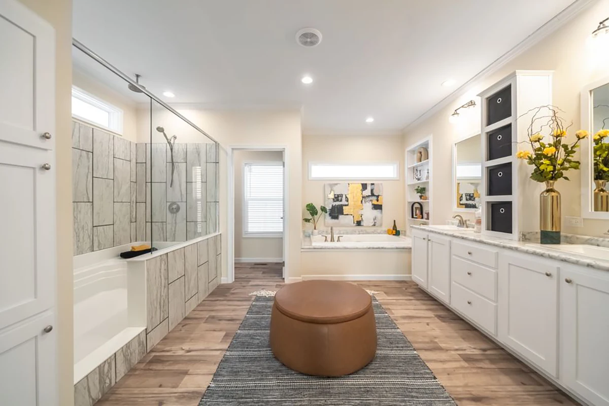 Spacious modern bathroom with a glass-enclosed shower, bathtub, dual sinks, white cabinets, a round ottoman, and decorative accents on countertops.