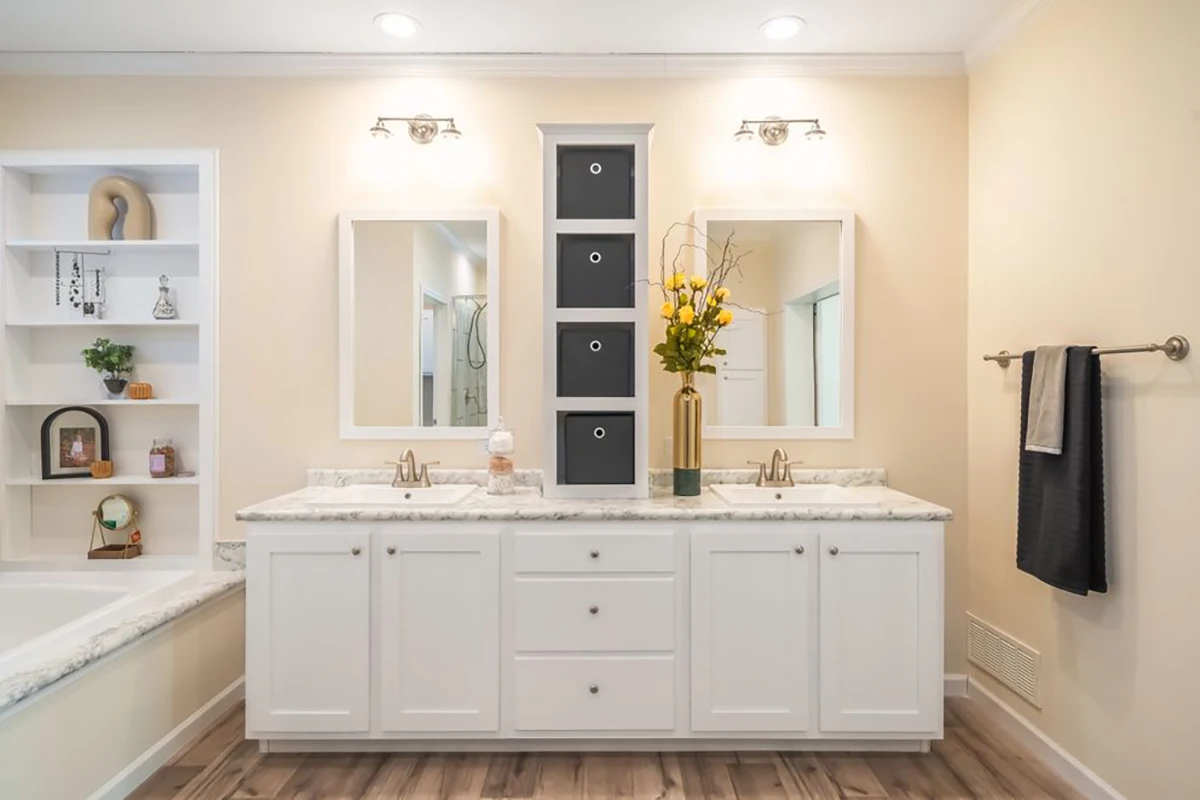 Bright bathroom with double sinks, white cabinets, two mirrors, a tall black drawer unit between them, and a vase with yellow flowers on the counter.