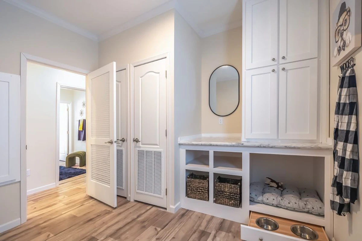 A mudroom with white cabinets, built-in bench, storage cubbies with baskets, a round mirror, hooks with hanging items, and pet beds and bowls on the floor.