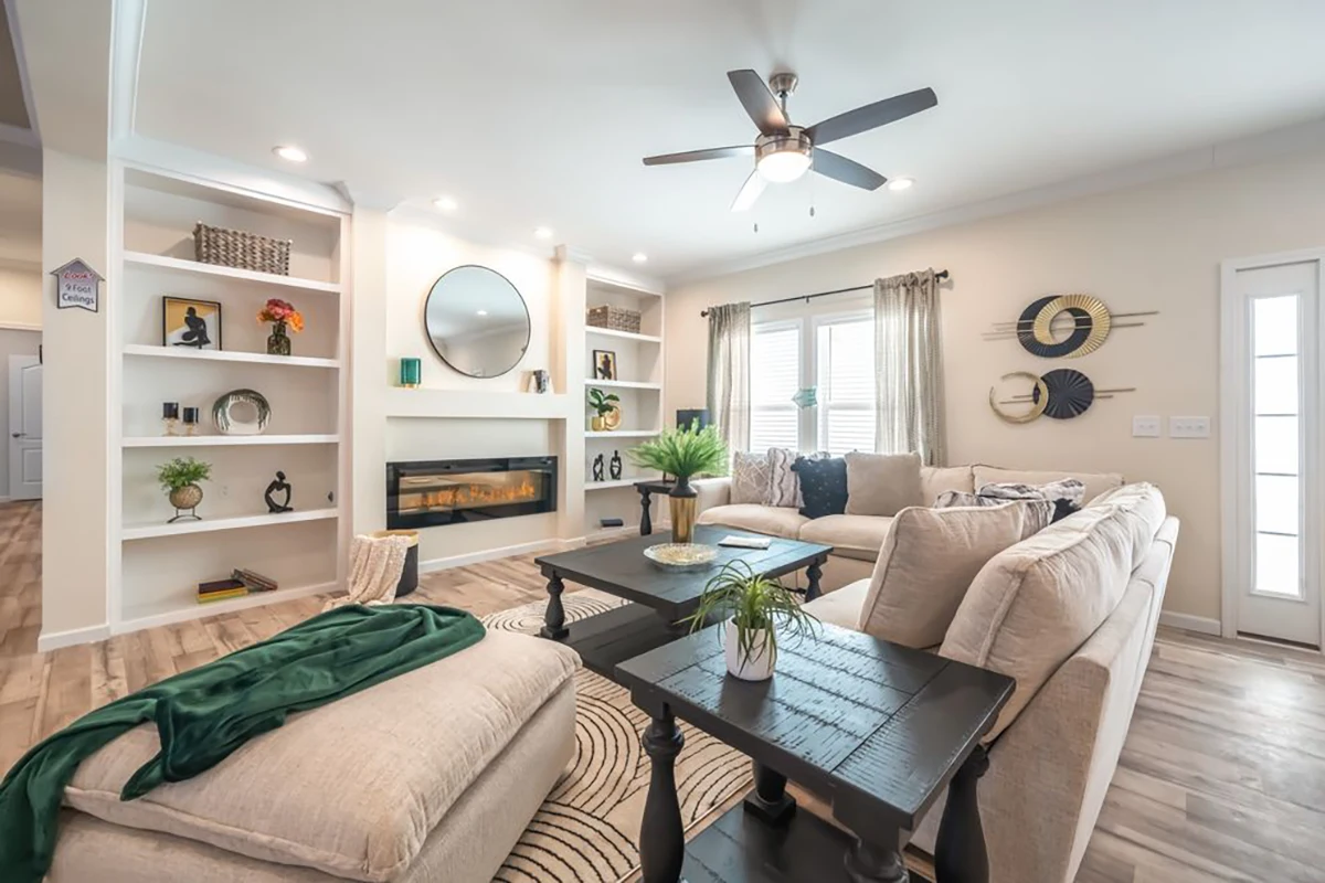 Modern living room with beige sofas, black coffee table, electric fireplace, wall shelves with decor, large windows with curtains, and ceiling fan.