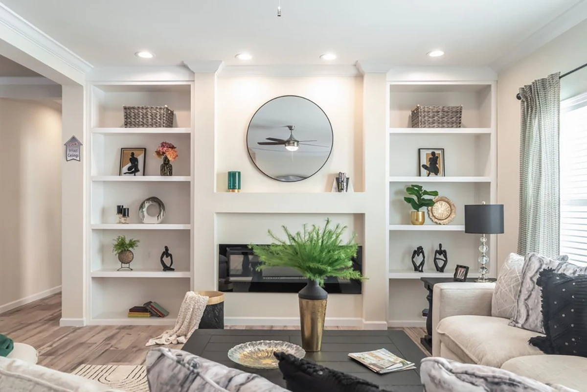 Modern living room with built-in white shelves, decorative items, a round mirror above a fireplace, neutral furniture, and a green plant on a black coffee table.