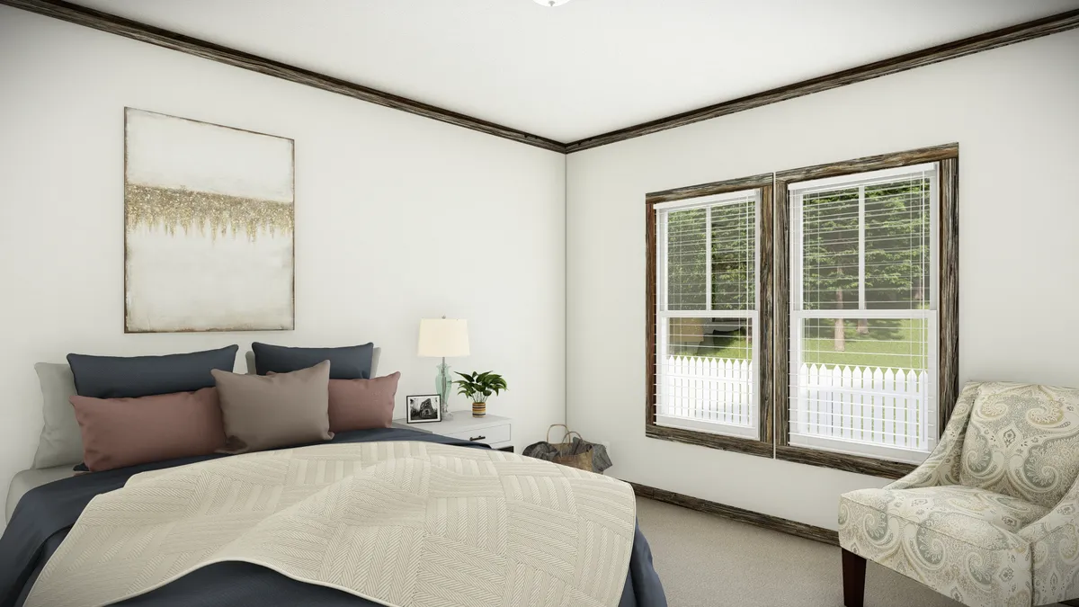 A bedroom with a bed, neutral bedding, pillows, a nightstand with a lamp, a window with blinds, a patterned chair, and a framed artwork on the wall.