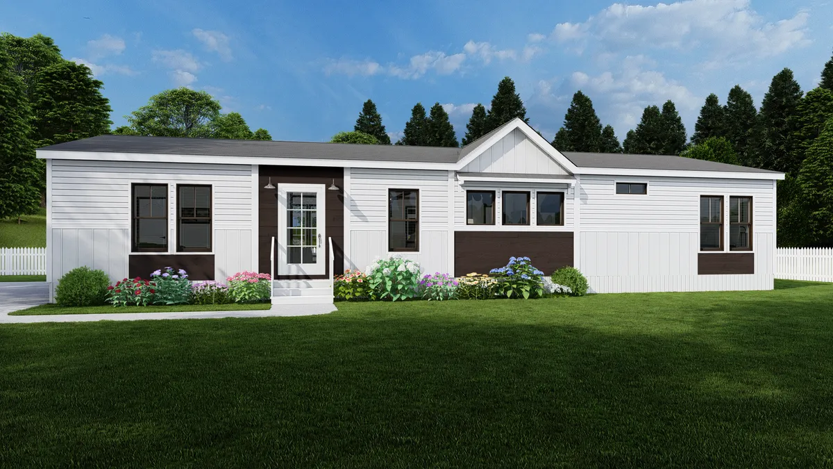 Single-story white manufactured home with dark trim, front door with steps, multiple windows, flower beds, grassy lawn, and trees in the background under a partly cloudy sky.