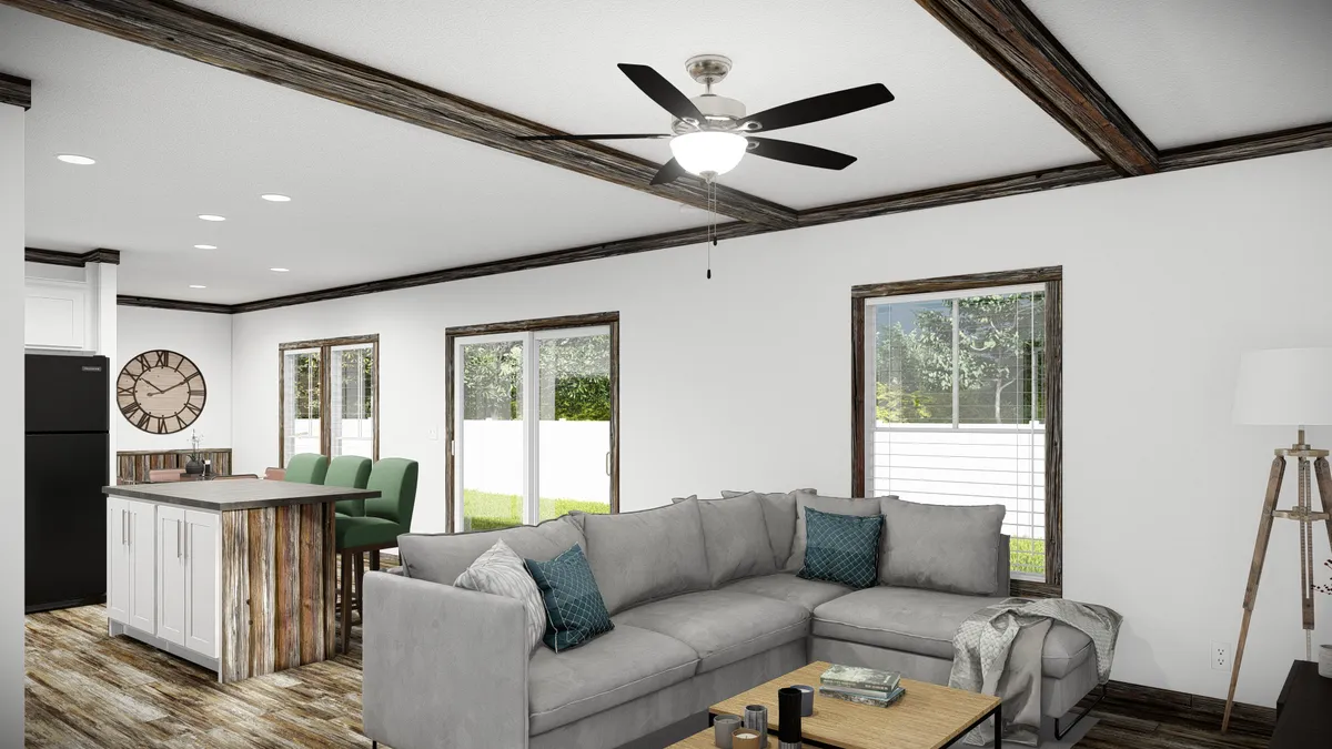 A modern living room with a gray sectional sofa, wooden coffee table, ceiling fan, and open kitchen area with green barstools and rustic wood accents. Large windows let in natural light.