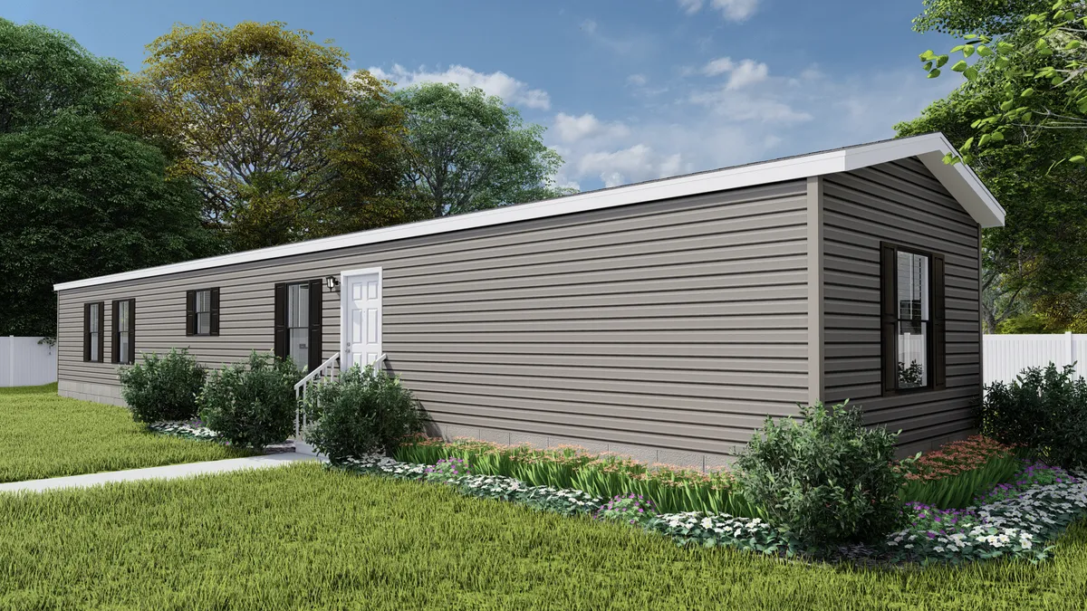 A single-story, gray manufactured home with a white door and windows, surrounded by green grass, shrubs, and trees under a partly cloudy sky.