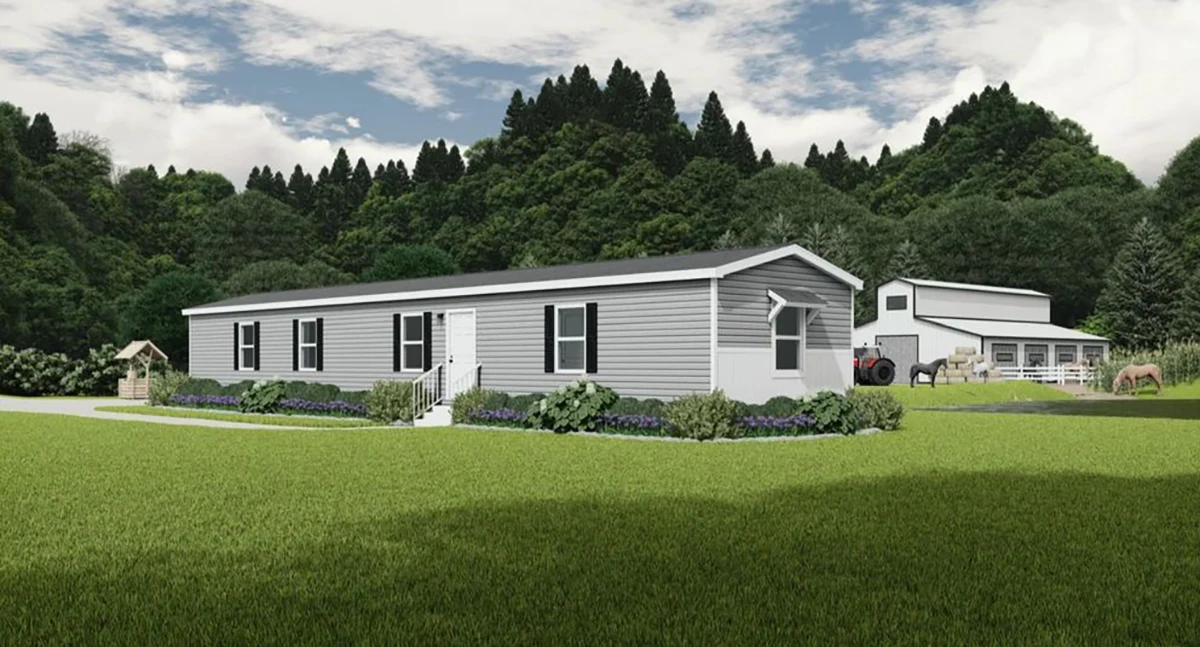A gray manufactured home with white trim sits on a green lawn, with trees, a barn, tractor, and horses in the background under a partly cloudy sky.
