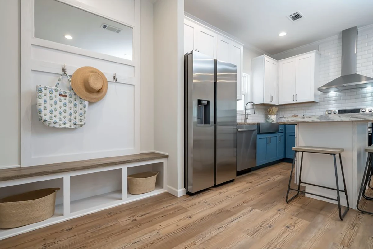 Modern kitchen and entryway with stainless steel fridge, white cabinets, blue farmhouse sink, wooden bench, hooks with a hat and tote, and two baskets on a wooden floor.