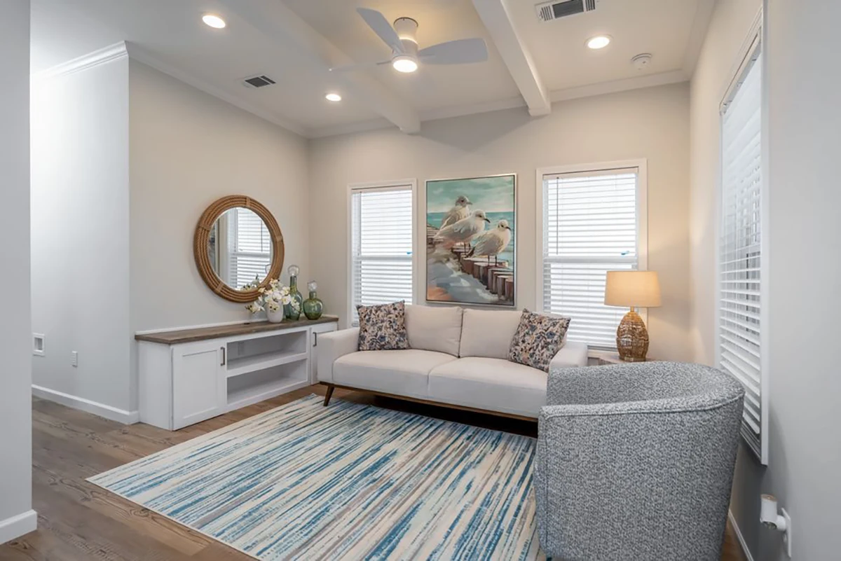 A modern living room with a beige sofa, blue-patterned rug, round mirror, gray armchair, and bird artwork above the couch, lit by natural light from three windows.