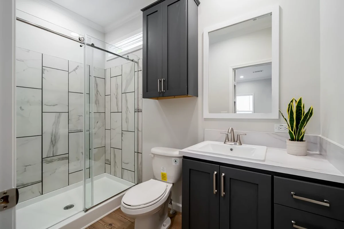 Modern bathroom with a glass-enclosed shower, gray cabinets, white sink, toilet, and a potted plant on the countertop.