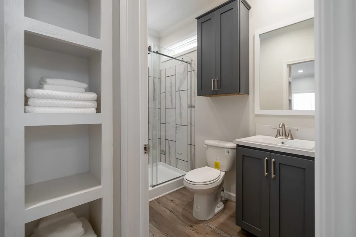 Modern bathroom with gray cabinets, white countertop, toilet, glass shower, and built-in shelves stocked with folded white towels. Wood-look flooring and neutral wall colors.