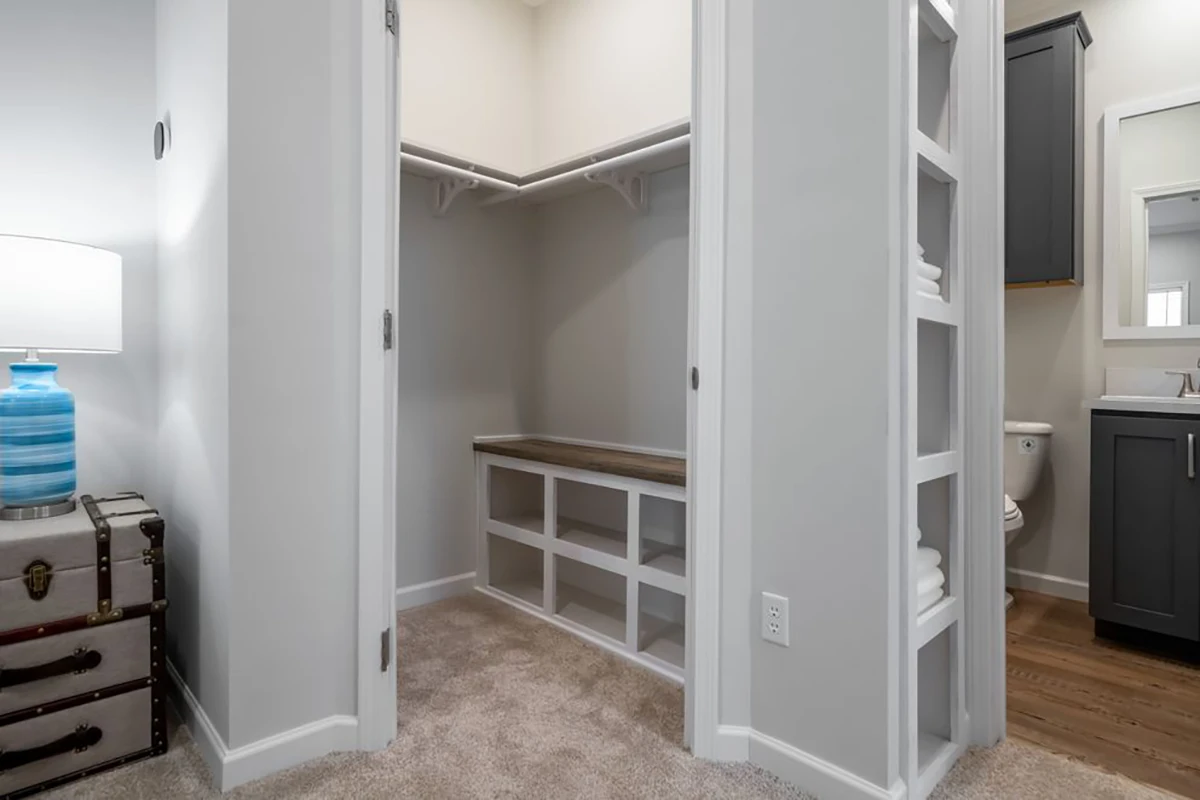 Open walk-in closet with white shelves and wooden bench, beige carpet flooring, and adjacent bathroom visible to the right.