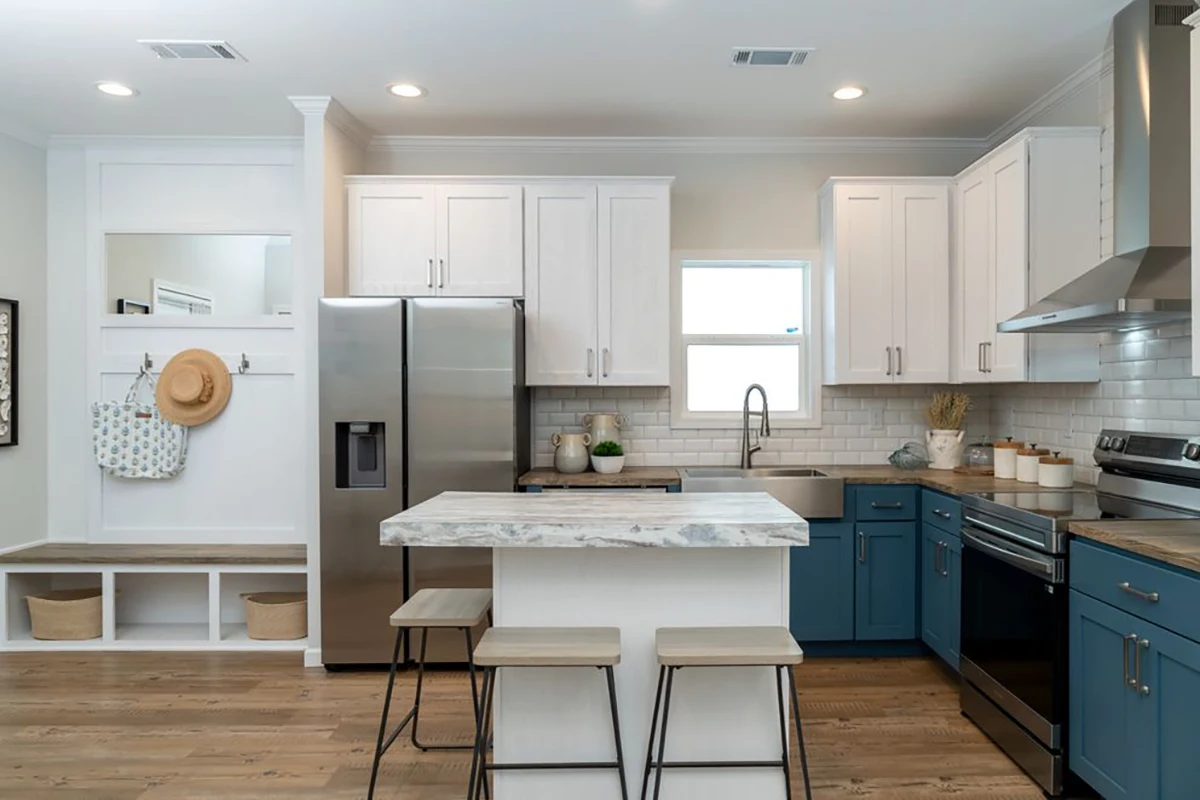 Modern kitchen with white upper cabinets, blue lower cabinets, stainless steel appliances, a center island with stools, and a small entryway with hooks and storage on the left.