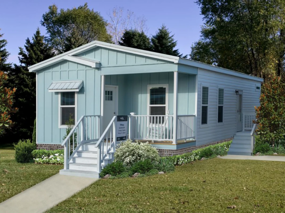 Light blue single-story manufactured home with white trim, a small porch with steps, surrounded by lawn, shrubs, and trees under a clear sky.