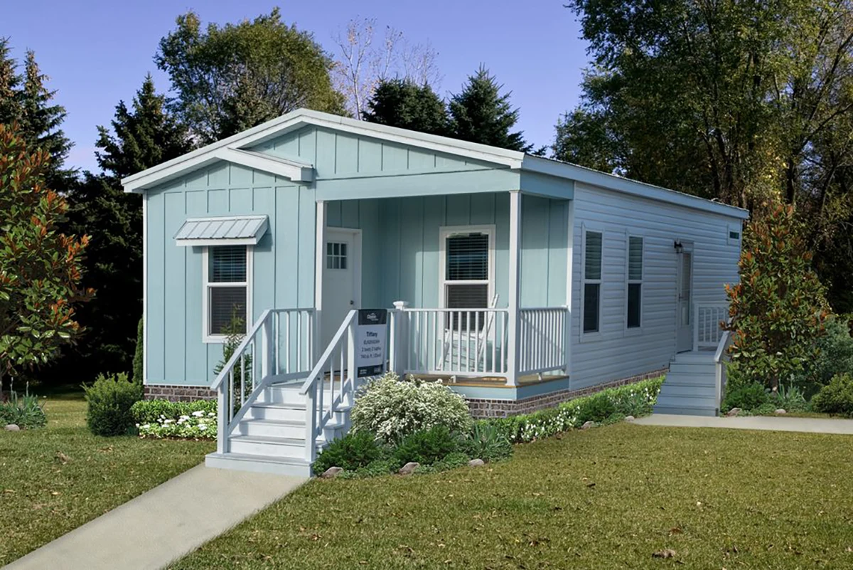 Light blue single-story manufactured home with white trim, a small porch with steps, surrounded by lawn, shrubs, and trees under a clear sky.