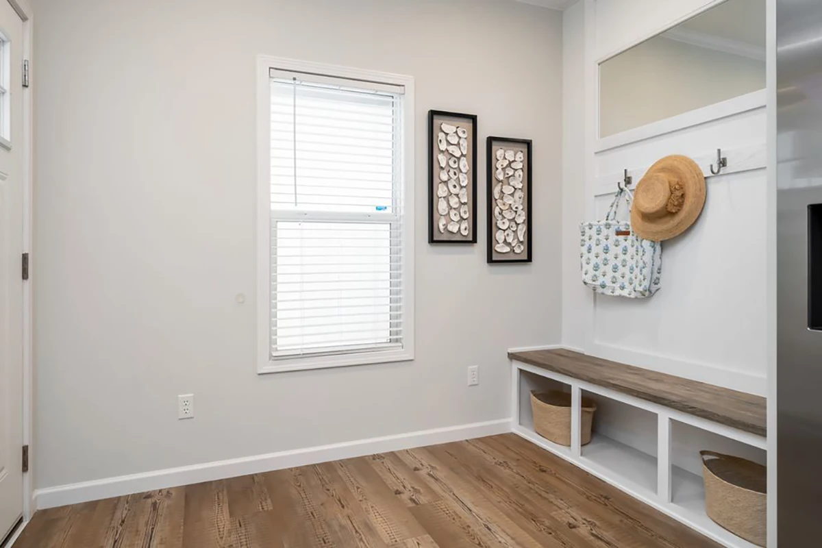 A mudroom with wooden bench seating, storage baskets below, hooks holding hats and a bag, wall art, a window with blinds, and light-colored walls and flooring.