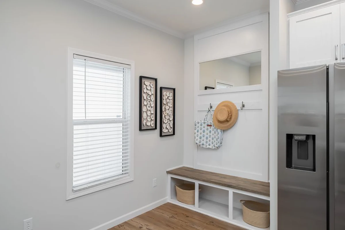 A mudroom with a wooden bench, storage baskets underneath, a wall shelf with hooks holding a hat and bag, two framed artworks, and a window with blinds.