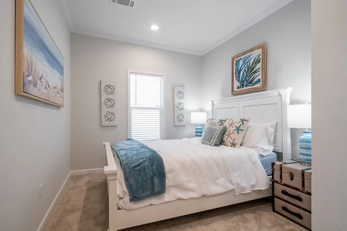 A neatly made bed with white and blue bedding is centered in a small bedroom with coastal-themed decor, two lamps, and a trunk used as a nightstand.