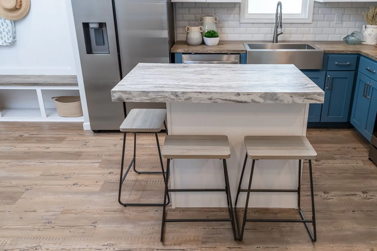 A small kitchen island with a gray marble countertop and two wooden stools is placed near blue cabinets and a stainless steel refrigerator.