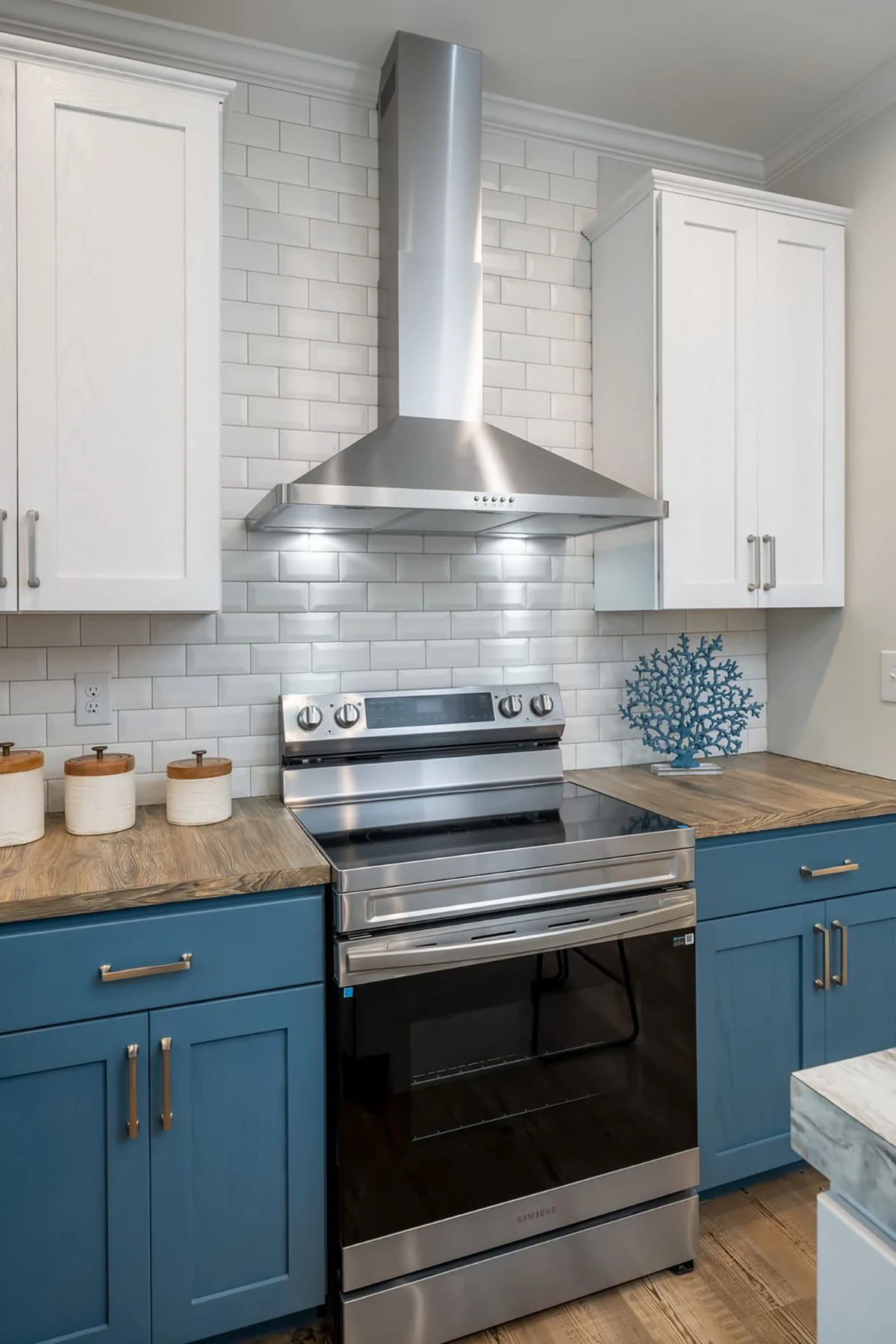 Modern kitchen with blue lower cabinets, white upper cabinets, a stainless steel stove and range hood, wood countertops, and white subway tile backsplash.