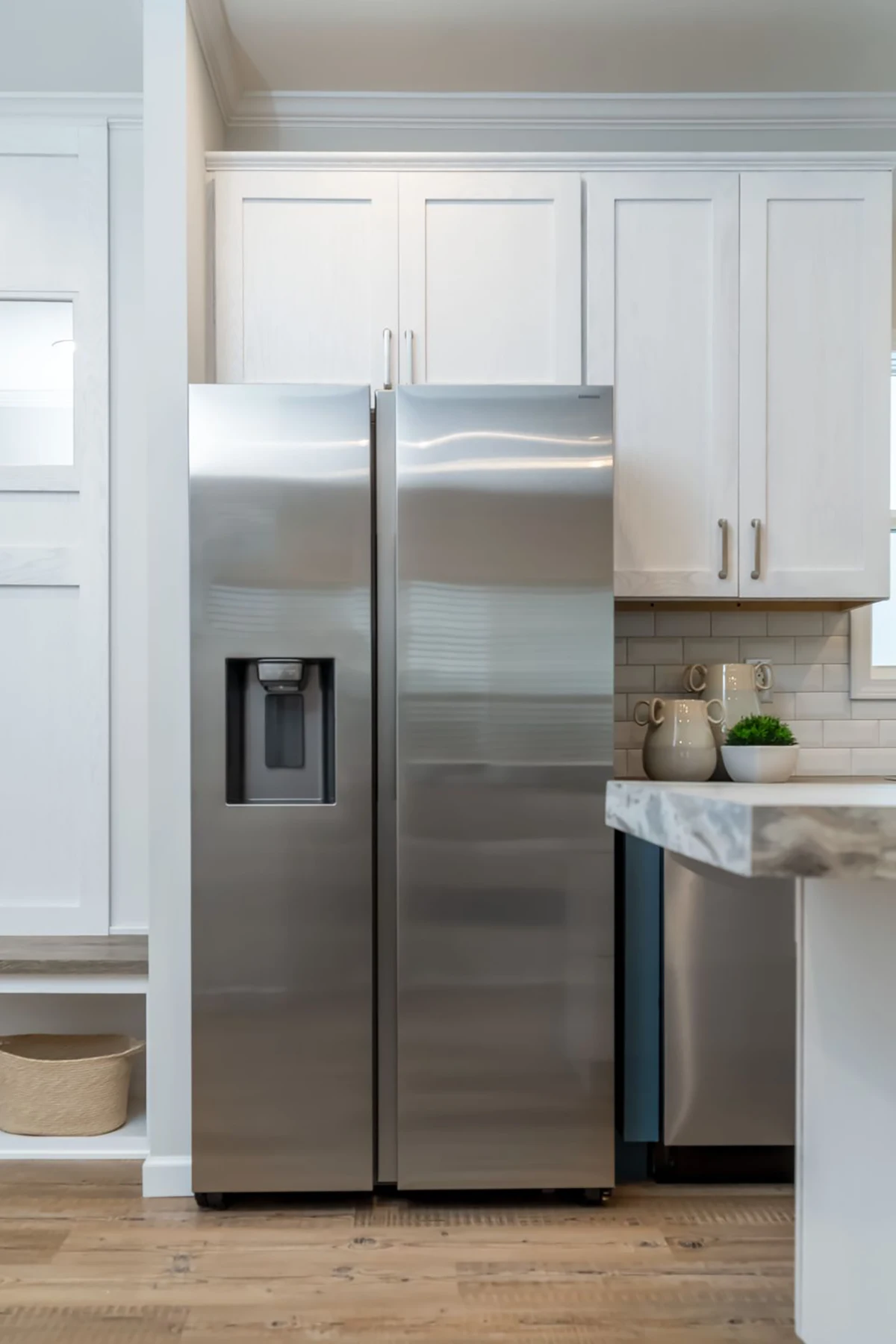 A modern stainless steel side-by-side refrigerator with a water and ice dispenser, set in a kitchen with white cabinets and wood flooring.