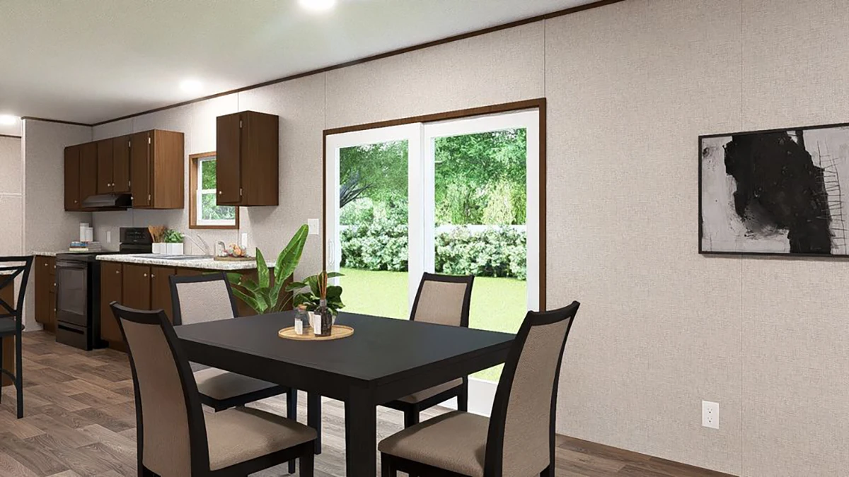 A dining area with a black table and four chairs is next to large glass doors; a kitchen with brown cabinets is in the background, and greenery is visible outside.