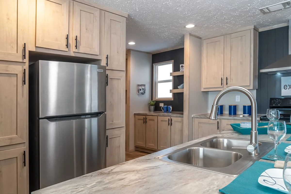 Modern kitchen with light wood cabinets, stainless steel refrigerator, double sink, marble countertop, and blue accents. Natural light enters through a window in the background.